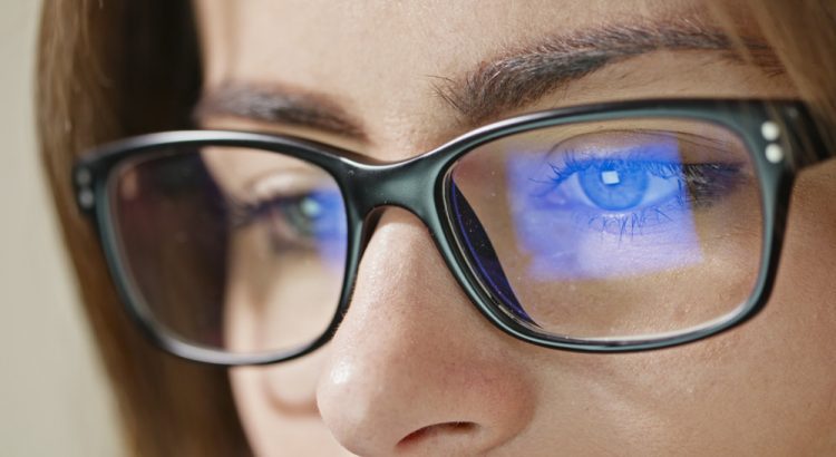 Mulher de óculos olha para o notebook. Na lente, a luz azul-violeta é refletida