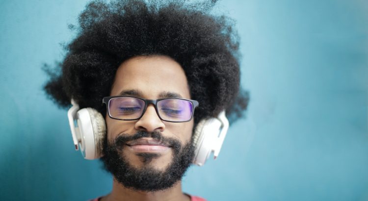 Homem aparentemente de Home Office, sentado em frente do computador usando óculos de grau e fones de ouvido. Sua expressão é de tranquilidade.