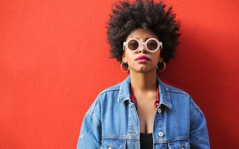 Mulher negra, de black power, encostada em uma parede vermelha, combina o óculo com a cor do cabelo