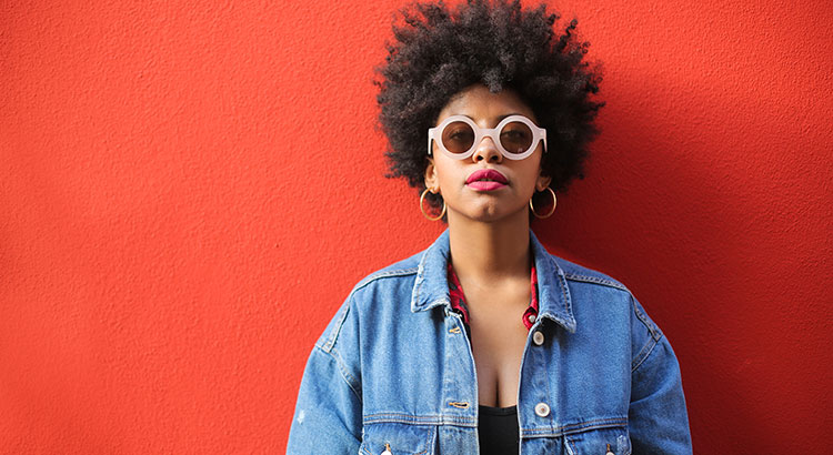 Mulher negra, de black power, encostada em uma parede vermelha, combina o óculo com a cor do cabelo