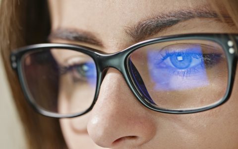 Mulher de óculos olha para o notebook. Na lente, a luz azul-violeta é refletida