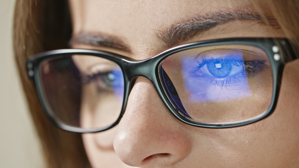 Mulher de óculos olha para o notebook. Na lente, a luz azul-violeta é refletida