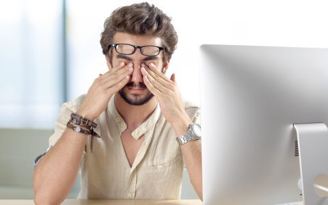 Homem leva as mãos na pálpebra tremendo, em frente ao computador