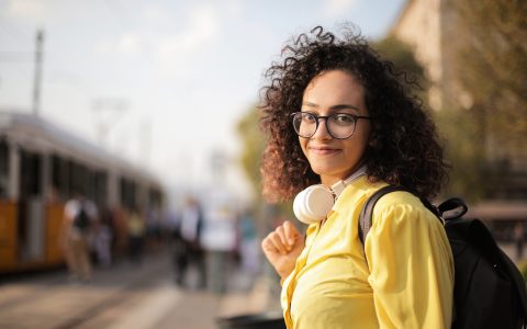 Modelo em um fundo urbano desfocado, olhando para a câmera, com uma camisa amarela e óculos no clima de outono