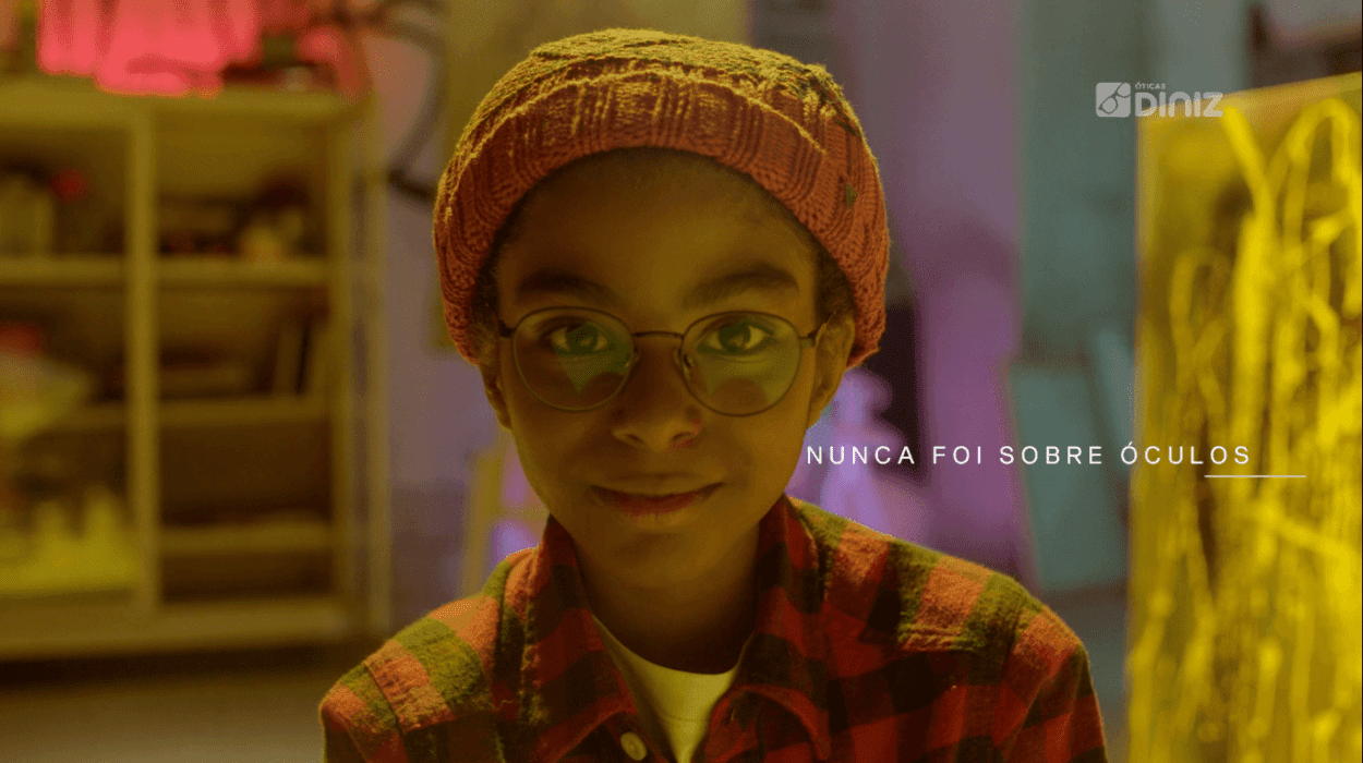 Menino negro de óculo, de blusa xadrez e touca olha para a câmera.