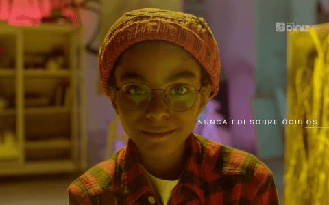 Menino negro de óculo, de blusa xadrez e touca olha para a câmera.