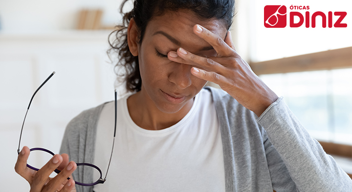 Mulher negra com a vista cansada segura os óculos com a mão direita e coça os olhos com a mão esquerda